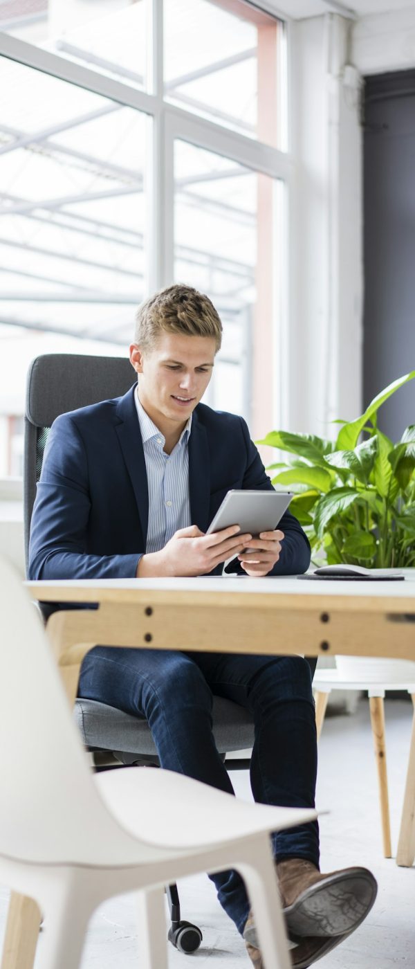 Confident young businessman sitting at desk in office using tablet